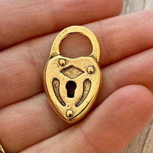 May include: A heart-shaped gold-toned metal padlock charm featuring a central keyhole, a diamond-shaped cutout above the keyhole, and three small circular decorative studs positioned around the perimeter of the lock face.
