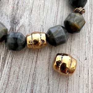 May include: A bracelet featuring faceted dark gray beads with a chatoyant sheen, interspersed with polished gold-tone metal beads. One additional gold-tone metal bead with a textured, organic surface rests separately near the bracelet strand.