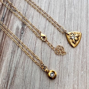 May include: Three gold-colored necklaces with different pendant designs. One pendant is a small, round shape with a central clear stone. Another is a shield shape with a floral design and a clear stone. The necklaces are on a wooden surface.