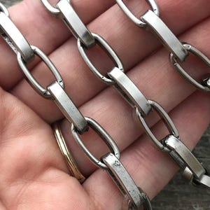 Puede incluir: Primer plano de una cadena de metal plateado con eslabones ovalados y barras rectangulares. La cadena se sostiene en una mano, mostrando el brillo metálico y la construcción detallada del componente de joyería.