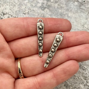 May include: Two silver-toned metal charms, each with a triangular shape and a loop at the top. The charms are adorned with a series of small, raised, round details. The charms are shown against a neutral background.