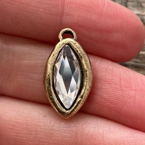 May include: A gold-toned metal charm with a clear, marquise-cut crystal center. The charm has a small loop at the top for attaching to jewelry. The crystal is faceted, reflecting light. The charm's frame has a textured, antique finish.