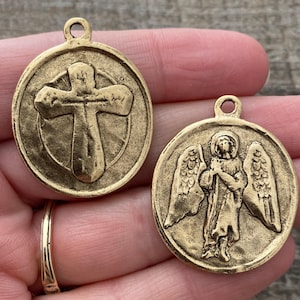 May include: Two antique gold-colored pendants. One pendant features a cross within a circle, the other depicts an angel with outstretched wings. Both pendants have a textured surface and a small loop at the top for hanging.