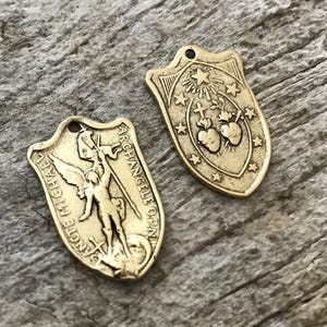 May include: Two antique gold-colored shield pendants. One pendant features an image of a figure with wings and the words "SANCTE MICHAEL ARCHANGELUS OPN". The other pendant has stars, crosses, and hearts.