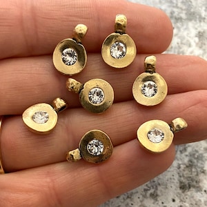 Puede incluir: Primer plano de varios colgantes pequeños de color dorado. Cada colgante tiene un diseño circular con una piedra transparente y brillante en el centro. Los colgantes están dispuestos sobre una superficie clara.