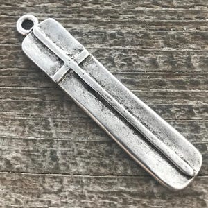 May include: A silver-colored pendant with a cross design. The pendant is rectangular with a loop at the top for hanging. The cross is centered on the pendant and has a slightly raised design.