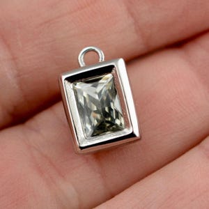 May include: A rectangular silver pendant with a clear, faceted gemstone. The pendant has a loop at the top for attaching to a chain or other jewelry. The gemstone is a light, neutral color, and the setting is polished silver.
