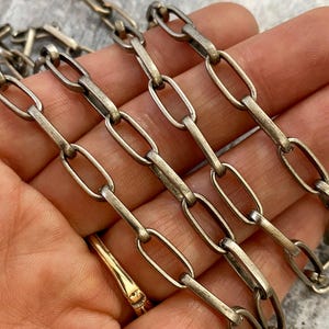 May include: Close-up of a silver-colored chain with elongated oval links. The chain is held in a hand, and a gold ring is visible. The chain appears to be vintage or antique, with a slightly tarnished finish.