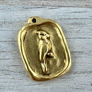 Puede incluir: Un colgante rectangular de metal dorado con bordes redondeados y superficie texturizada. El centro presenta un relieve de un pájaro posado en una rama. Un pequeño orificio circular está situado en la parte superior para sujetarlo.