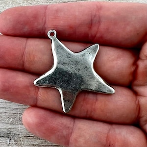 May include: A silver-colored star-shaped charm with a loop at the top for hanging. The charm has a distressed, vintage appearance, with visible texture and slight discoloration. The star's points are slightly rounded, and the metal has a dull sheen.