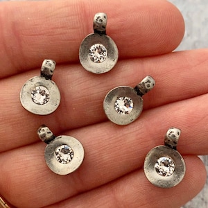 May include: Five small, silver-toned pendants with a circular design and a clear, sparkling crystal in the center. Each pendant has a small loop at the top. The pendants are displayed on a light-colored surface.