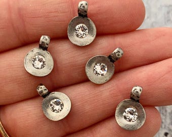 May include: Five small, silver-toned pendants with a circular design and a clear, sparkling crystal in the center. Each pendant has a small loop at the top. The pendants are displayed on a light-colored surface.