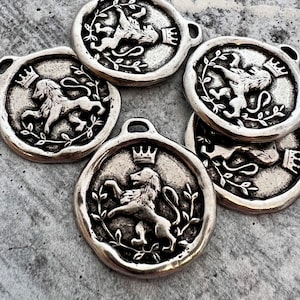 May include: A collection of circular silver-toned metal charms, each featuring a raised relief design of a lion wearing a crown. The lion is depicted in a rampant stance, surrounded by a delicate leafy branch motif. Each charm includes a small loop at the top for attachment, with a weathered, antiqued finish that highlights the intricate details of the embossed heraldic imagery.