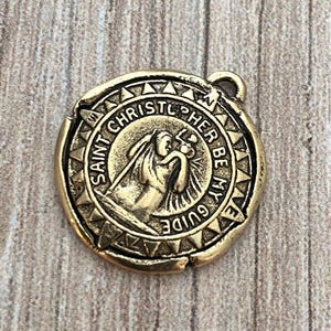 Op de afbeelding: Een ronde, antiek goudkleurige Sint-Christoffelmedaille. De medaille toont een afbeelding van Sint-Christoffel die een kind draagt, omringd door de woorden "Saint Christopher Be My Guide". De medaille heeft een lus aan de bovenkant om aan een ketting te bevestigen.
