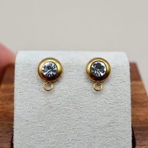 May include: A pair of gold-toned circular earring components featuring a central clear, faceted crystal stone and a small, integrated jump ring at the bottom for attaching additional jewelry elements or charms to the base of the setting.