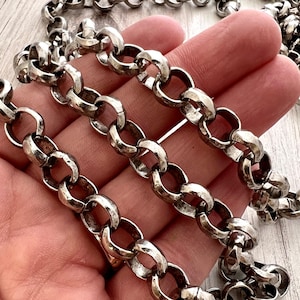 May include: Close-up of a silver-toned chain necklace with large, textured oval links. The chain is displayed against a neutral background, highlighting its metallic sheen and intricate design. The chain is a jewelry item.