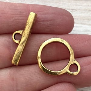 May include: A gold-toned toggle clasp set consisting of a bar component with a central loop and a circular ring component with an attached side loop. Both pieces feature a textured, matte finish and are designed for jewelry making and fastening.
