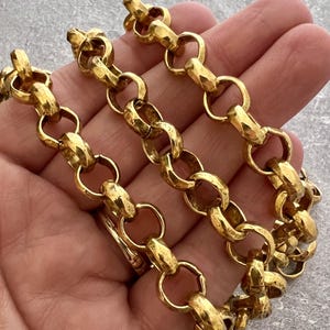 Puede incluir: Primer plano de un collar de cadena de color dorado. Los eslabones de la cadena son de forma ovalada y están conectados por anillos redondos. El collar tiene una superficie texturizada y un acabado brillante. La joya se muestra sobre un fondo neutro.