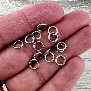 May include: Close-up of small, silver-colored jump rings. Some rings are open, while others are closed and interlocked. The rings are scattered across a surface, suggesting their use in jewelry making or crafts.