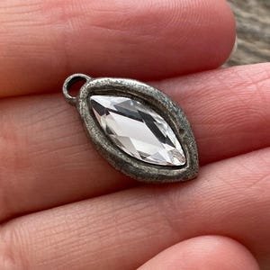 May include: A small, antique-style pendant with a clear, marquise-cut crystal set in a silver-toned metal frame. The frame has a textured, aged appearance, and a small loop is at the top for attaching to a chain or other jewelry component.