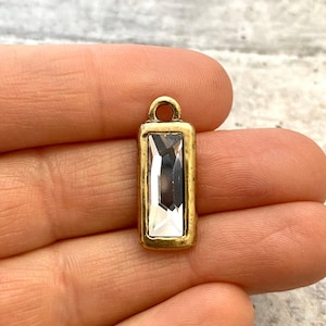 May include: A rectangular pendant charm featuring a clear, faceted baguette-cut crystal set within a textured, antique-gold tone metal frame. The charm includes a small circular loop at the top for attachment, showcasing a metallic finish.