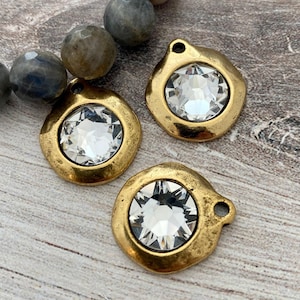 May include: Three gold-toned pendants with a clear, faceted crystal center. Each pendant has a unique, irregular shape and a small loop at the top. The pendants are displayed on a light-colored, textured surface, with a strand of gray beads in the background.