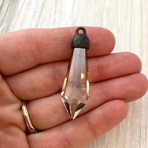 May include: A clear, faceted crystal pendant with a dark metal cap and loop. The pendant is held in a hand, showcasing its pointed, elongated shape and light refraction. The crystal is approximately 2 inches long.