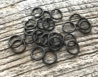 May include: A collection of small, circular metal jump rings with a dark, matte charcoal finish. Each ring features a single, clean cut opening, allowing them to be easily pried apart and closed for jewelry making or various craft assembly projects.