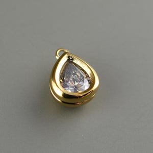 May include: A gold-toned teardrop-shaped pendant featuring a clear, faceted stone set within a smooth, polished metal frame. The pendant includes a small circular jump ring at the top for attachment, showcasing a reflective and metallic finish.