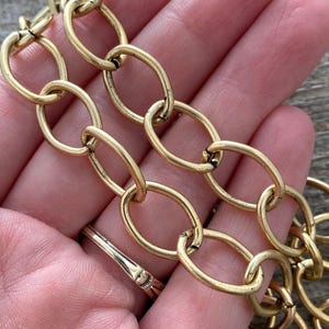 May include: Close-up of a gold-colored chain with oval links. The chain is held in a hand, and a gold ring is visible on a finger. The chain has a vintage or antique appearance.