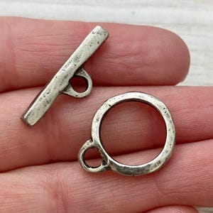 Peut inclure: Gros plan sur un ensemble de fermoirs à bascule argentés. L'ensemble comprend une barre rectangulaire avec une boucle et un anneau circulaire avec une boucle. Le métal a un aspect légèrement vieilli, suggérant un style antique ou vintage. Idéal pour la création de bijoux.