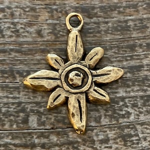 Pode incluir: Um pingente de metal cor de latão em forma de flor de edelweiss com um detalhe circular central e oito pétalas. A superfície tem um acabamento antigo e desgastado com áreas escuras rebaixadas e um pequeno elo no topo para fixação.