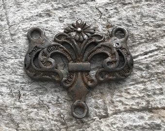 Könnte beinhalten: Ein dunkles, verwittertes Metallverbindungsstück mit einem verzierten, symmetrischen Schnörkeldesign und einem zentralen Blumenmotiv an der Oberseite. Das Bauteil verfügt über drei kreisförmige Ösen zur Befestigung, je eine an den oberen Ecken und eine an der Basis.