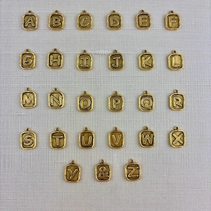 Könnte beinhalten: Eine Sammlung goldfarbener Alphabet-Anhänger, jeweils in einem quadratischen Rahmen. Die Buchstaben A bis Z sowie ein kaufmännisches Und werden angezeigt. Die Anhänger sind auf einer weißen, strukturierten Oberfläche angeordnet.