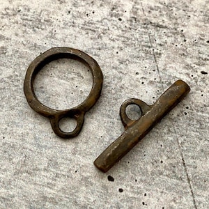 May include: A set of two antique bronze-colored metal toggle clasps. One is a ring with a loop, and the other is a bar with a loop. The clasps have a rustic, aged appearance, suggesting they are vintage or handcrafted.