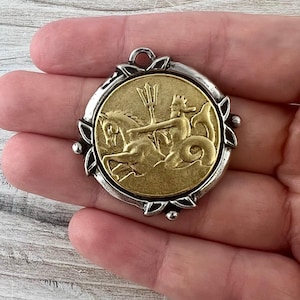Puede incluir: Un colgante redondo con una moneda dorada que representa una figura montada en un caballito de mar, enmarcada por un borde decorativo plateado. El colgante tiene un lazo en la parte superior para colgar.
