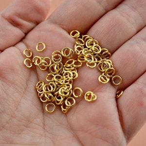 May include: A collection of small, gold-colored jump rings, scattered across a person's open palm. The rings are circular and appear to be made of metal, likely for jewelry making or crafting projects. The lighting highlights the metallic sheen.