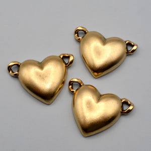 May include: Three gold-toned metal heart-shaped charms, each featuring a smooth, rounded surface and two small loops on opposite sides for attachment. The hearts have a matte finish and a uniform, solid appearance, arranged in a cluster.