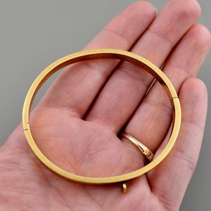 May include: A gold-colored bangle bracelet with a hinged clasp and a small loop. The bracelet is circular and appears to be made of metal. The bracelet is held in a hand against a neutral background.