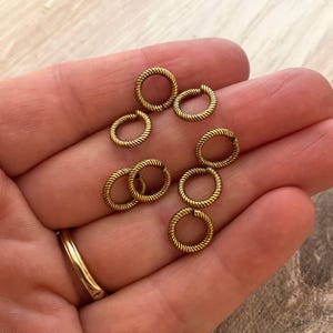 Könnte beinhalten: Eine Sammlung kleiner, goldfarbener Sprungringe mit einer gedrehten Seiltextur. Die Ringe sind offen und bereit für die Verwendung in der Schmuckherstellung oder anderen Bastelarbeiten. Ein goldener Ring ist in der linken unteren Ecke sichtbar.