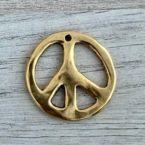 May include: A gold-coloured peace sign pendant. The pendant is round with a classic peace symbol design. It has a small hole at the top for stringing. The pendant is made of a shiny, metallic material.