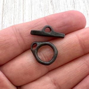 Puede incluir: Dos pequeños cierres de metal oscuro. Uno tiene forma de anillo y el otro es una barra con un lazo. Los cierres son probablemente para joyas o pequeños accesorios. Los cierres se sostienen en la mano de una persona.