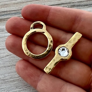 May include: Two gold-colored toggle clasps. One is a ring with a loop, the other is a bar with a round, clear crystal. The clasps have a hammered texture. These clasps are used for jewelry making.