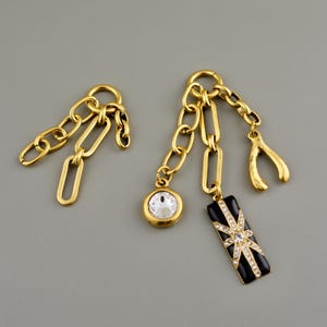 May include: Gold-toned chain earrings with various charms. One earring features a clear crystal pendant and a black rectangular charm with a starburst design. The other earring has a chain design. Jewelry.