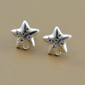 May include: A pair of silver-toned star-shaped earrings. Each earring features five smaller star-shaped embellishments, each set with small, clear gemstones. A small loop is attached to the bottom of each earring.