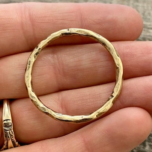 May include: A circular gold-toned ring featuring a distinct hammered texture across its entire surface. The metal band has an uneven, organic finish with small indentations that catch the light, creating a reflective and textured appearance.