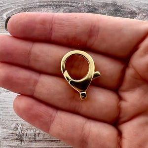 May include: A gold-colored swivel clasp, featuring a circular ring and a spring-loaded lever for secure closure. The clasp is designed for attaching to bags, keychains, or other accessories. The metal has a shiny finish.