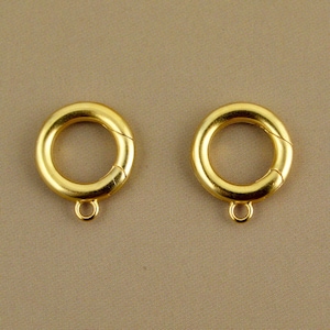 May include: Two gold-colored, circular clasps with a spring-ring closure and a small loop at the bottom. The clasps are set against a neutral, tan background. These jewelry findings are designed for necklaces, bracelets, or other jewelry.