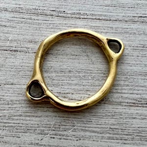 Puede incluir: Un componente conector de metal en tono dorado que presenta un anillo circular central con dos bucles circulares integrados más pequeños que se extienden desde los lados opuestos. La superficie tiene un acabado liso y pulido con una forma orgánica ligeramente irregular.