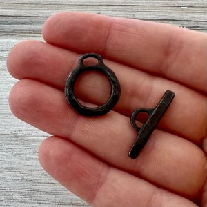 May include: Two dark bronze metal jewellery clasps. One is a ring with a loop, and the other is a bar with a loop. The clasps are likely for necklaces or bracelets. The clasps are shown against a neutral background.